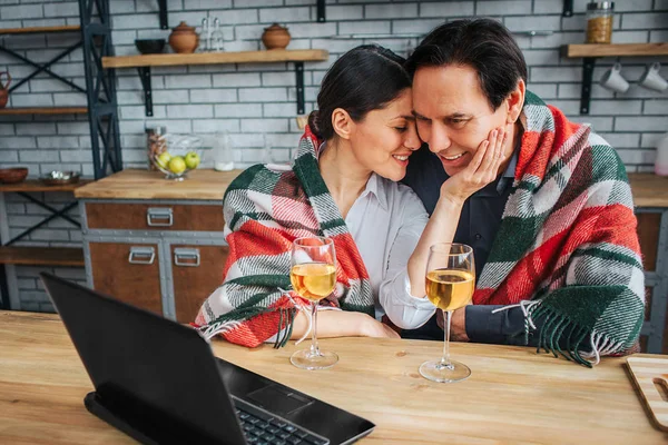 Womn sit with man at table in kitchen. She hold her hand on his cheek and lean to him. Man look on laptop. They have glasses of wine on table. — Stock Photo, Image