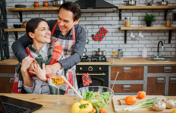 Nice couple is in kitchen. Guy stand behind woman and embrace her. She has blanket on shoulders. Woman sit at table and hold husband hands in her. — Φωτογραφία Αρχείου