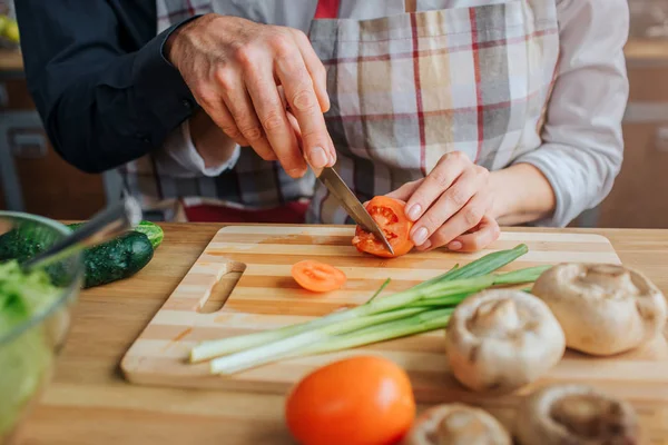 Skär syn på man och kvinna händer som håller kniven och skära tomat vid bord. Han hjälper henne. Det finns grön lök, svamp och skål med letuce på bordet. — Stockfoto