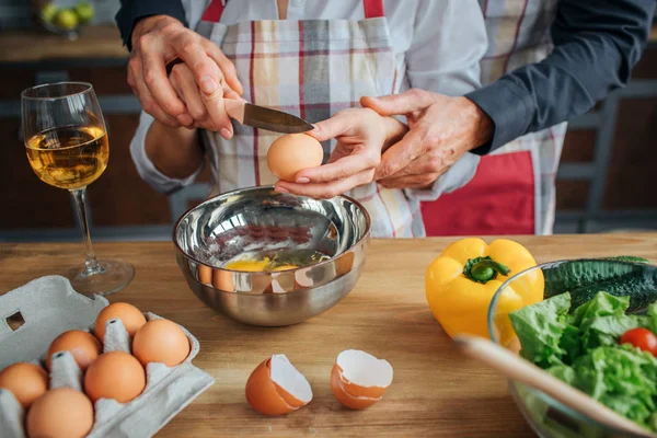 Man händer att hjälpa kvinnan att bryta ägg med kniv. Han står bakom henne i köket. Det finns ingredienser sallad och glas på vin på bordet. — Stockfoto