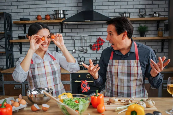 Funny young woman sit at table with husband in kitchen. She covered eyes with tomatoes. Man amazed and wondered. He look on her. — Stock Photo, Image