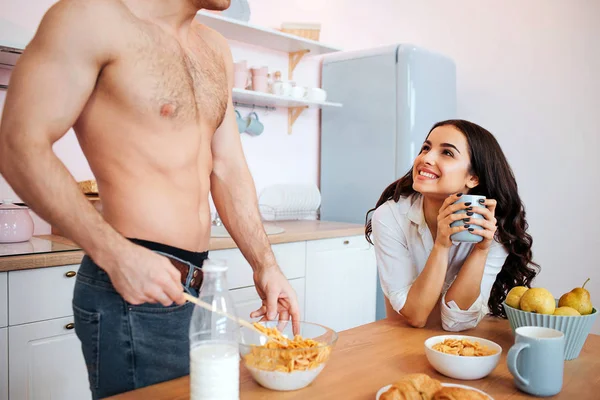 Corte vista de sexy mens corpo na cozinha. O tipo fica à mesa e mistura leite com flocos de milho. Jovem alegre olhar para ele e sorrir. Ela segura o copo . — Fotografia de Stock