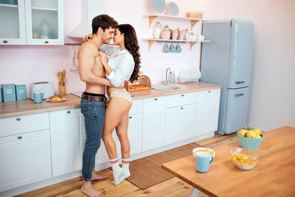 Alegre casal jovem sexy na cozinha. Abraçam-se e olham um para o outro. Sorriem e posam. Pequeno-almoço esperar na mesa . — Fotografia de Stock