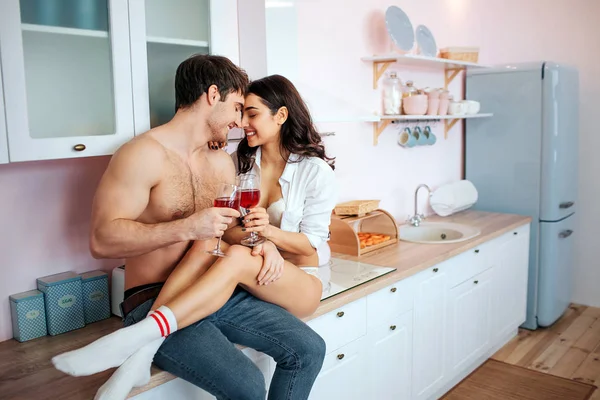 Jovem casal alegre e feliz na cozinha. Sentam-se no armário da cozinha e sorriem. As pessoas têm copos de vinho tinto nas mãos . — Fotografia de Stock