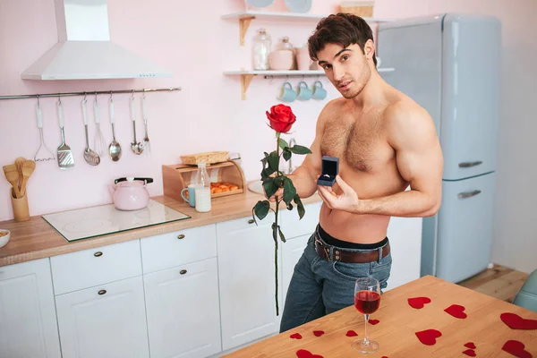 Ωραίος και paassionate νεαρός άνδρας σταθεί στην κουζίνα στο τραπέζι. Κρατούσε την κόκκινο τριαντάφυλλο και το κουτί με το δαχτυλίδι. Νεαρός άνδρας στάση και θέτουν στην κάμερα. — Φωτογραφία Αρχείου