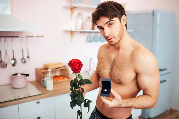 Beau jeune homme swtand nu dans la cuisine. Il regarde et pose devant la caméra. Guy tenir rose rouge et boîte avec anneau dans les mains . — Photo
