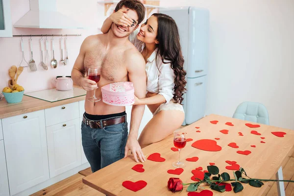 Cheerful sexy young woman stand with man in kitchen. She hold box with present. Mans eyes are closed with her hand. Guy has glass of red wine. — ストック写真