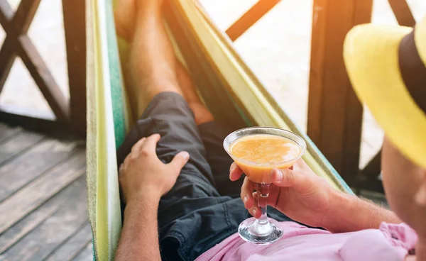 Schöner junger Mann, der in einer Hängematte an einem sonnigen Strand am Meer liegt und Cocktails in Händen hält. Sonne scheint hell. — Stockfoto