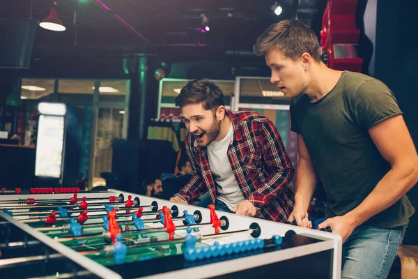 Jeunes hommes heureux excités jouant au baby-foot dans la chambre. Ils se tiennent ensemble et regardent le jeu . — Photo