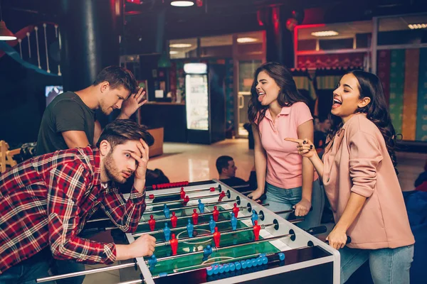 Happy young women look at men and shrink. They win game. Guys lost it. They dissapointed. Young people stand in playing room. — Stock Photo, Image