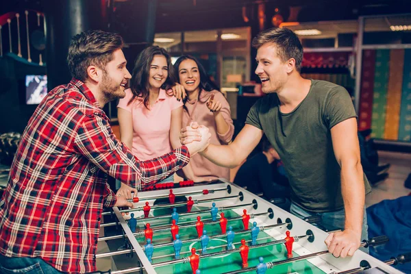 Happy young men stand at table soccer in playing room and hold each other hands. They have competition. Young women stand behind and look at them.