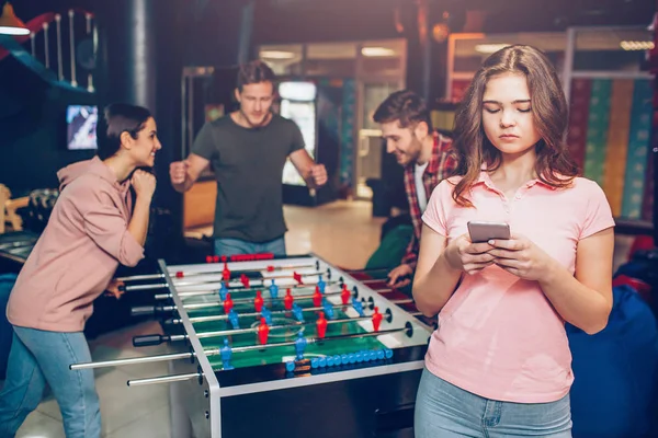 Jeune modèle en chemise rose tenir le téléphone dans les mains. Joyeux jeune équipe jouant au baby-foot dans la salle de jeu. Guy debout vers la femme . — Photo