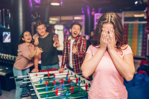 Жорстокі молоді люди стоять за столом футболу в ігровій кімнаті і сміються. Вони вказують на жінку в рожевій сорочці. Вона прикриває обличчя руками . — стокове фото