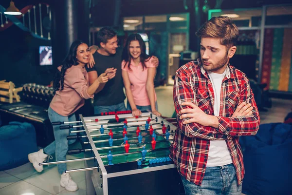 Upset young man stand in front in playing room. He look backwards. His friends standing behind and laughing. They cheerless. — Stock Photo, Image