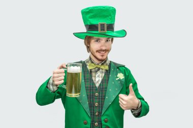 Yeşil saint Patrick takım elbise holding kupa bira ve gülümseme mutlu delikanlı resmi. O büyük başparmak bekle. Gri arka plan üzerinde izole.