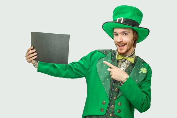 Joven feliz con traje verde sosteniendo un pedazo de papel negro. Lleva traje de santo Patricks. Aislado sobre fondo gris . — Foto de Stock