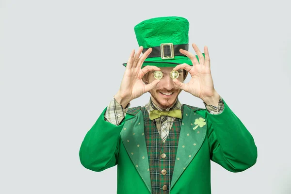 Boldog, gazdag fiatal ember, zöld színű tartsa két aranyat szemei előtt. Mosolyogsz, és örül. Guy zöld Szent Patricks jelmez viselnek. Elszigetelt, a szürke háttér. — Stock Fotó