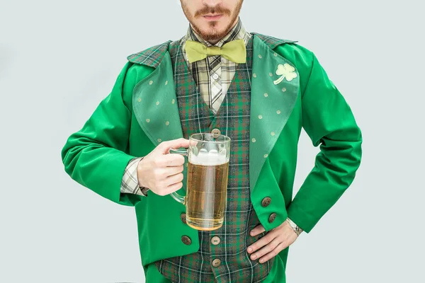 Vista de corte de hombre joven en traje verde sosteniendo taza de cerveza. Tiene otra mano en las caderas. El tipo usa traje de santo Patricks. Aislado sobre fondo gris . — Foto de Stock