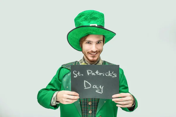 Jovem feliz em terno verde segurando tablet escuro com palavras escritas dia St. Patricks. O tipo olha para a câmara e sorri. Isolado sobre fundo cinzento . — Fotografia de Stock