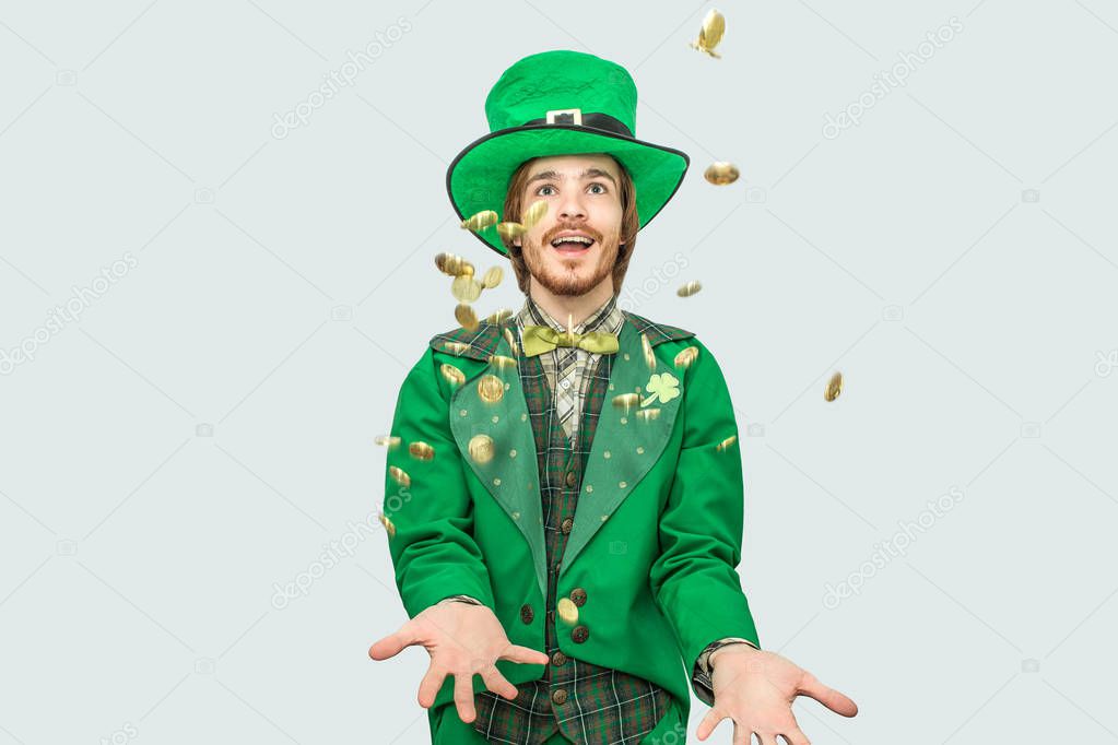 Happy rich young man in saint Patricks suit spreading golden coins up and smiling. They fly. Guy look up. Isolated on grey background.