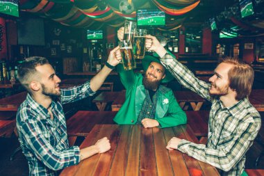 Barda bira kupaları birlikte tablo yukarıda tutan üç mutlu genç arkadaş. İnsanlar gülümse. Adam orta aşınmaya St. Patrciks yeşil uygun.