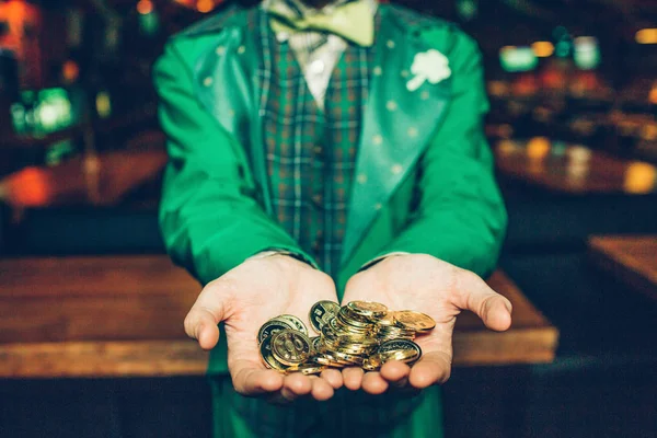Vista corte de cara em terno verde ficar em pub e segurar moedas de ouro nas mãos. Ele mostra-os à câmara. . — Fotografia de Stock