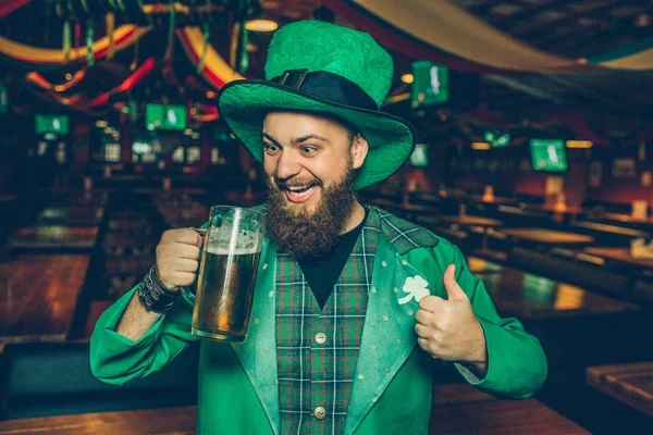 Happy young man in St. Patricks suit stand in pub and look at mug of beer. He hold big thumb up. Young man looks happy and smile.