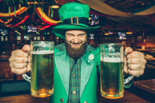 Jovem feliz e descontraído de St. Patricks só no pub. Ele segura duas canecas de cerveja e olha para a câmara. . — Fotografia de Stock