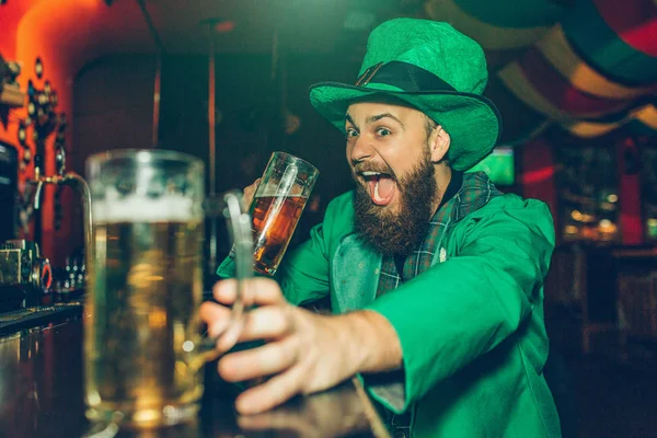 Glücklich aufgeregter junger Mann in St. Patricks grünem Anzug sitzt an der Theke in der Kneipe. er trinkt Bier aus einem Becher und greift nach einem anderen. — Stockfoto
