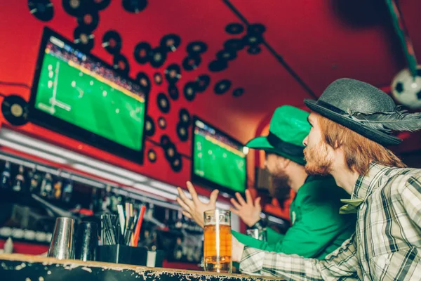 Two friends sit at bar counter in pub and watch football game. They serious and concentrated. One of them wear green St. Patricks suit. Mug of beer stand at bar counter. — Stock Photo, Image
