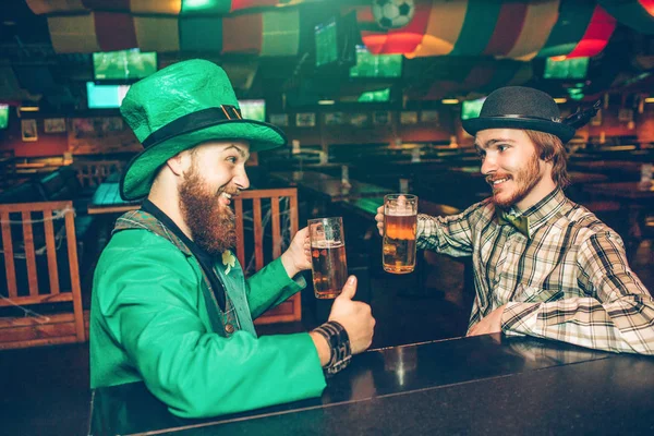 Cheerful happy young men sit in front of each other at bar counter in pub. They cheer and hold mugs of beer. Guy on left wear St. Patricks green suit.