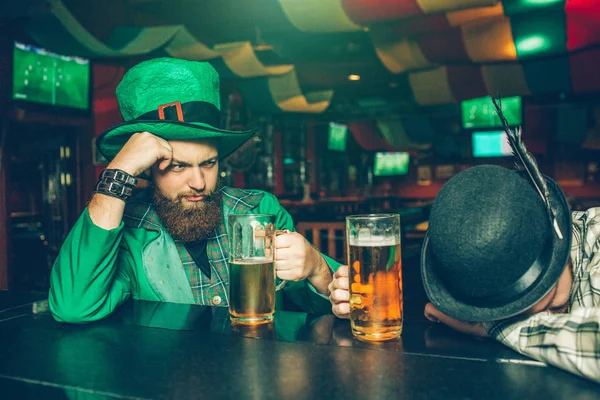 Drunk young man in green suit sit at bar counter in pub with friend. Another guy fell asleep. They have mugs of beer. — Stock Photo, Image