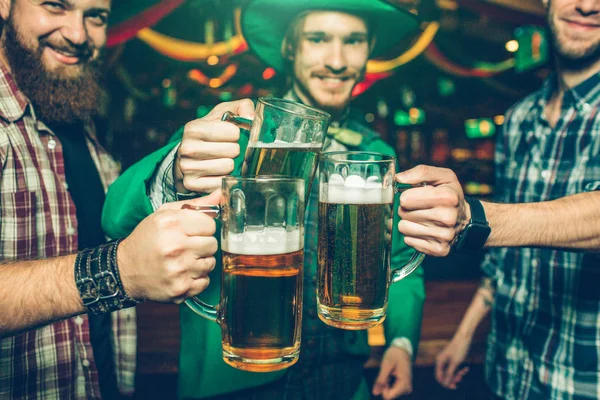 Glada lyckliga vänner står tillsammans i pub och heja. De håller muggar öl nära varandra. Människor ler. De bära festliga nära och grön kostym. — Stockfoto