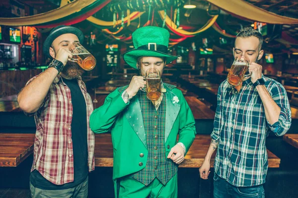 Három fiatal férfi képe állvánnyal együtt kocsmában, és inni sört, a bögre. Koncentrált. Fickó St. Patricks megfeleljen középső kopás. — Stock Fotó