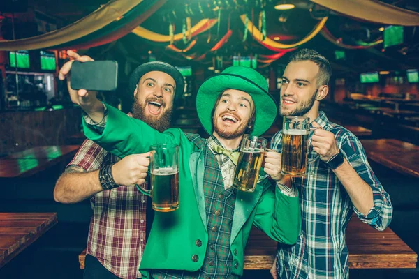 Glückliche junge Männer beim gemeinsamen Selfie in der Kneipe. Mann in der Mitte trägt St. Patricks Anzug und hält Telefon in den Händen. sie lächeln. — Stockfoto