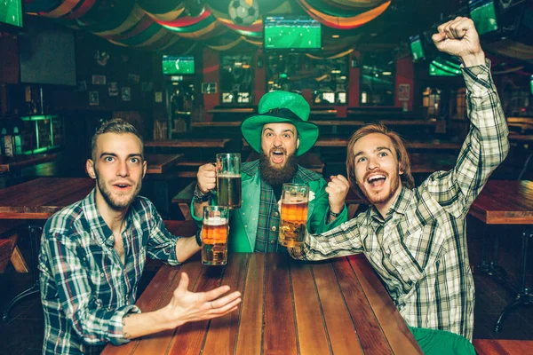 Aufgeregte junge Männer sitzen am Tisch in der Kneipe und freuen sich. Sie jubeln. Jungs halten Bierkrüge in den Händen. junger Mann in der Mitte trägt grünen St. Patricks Anzug. — Stockfoto