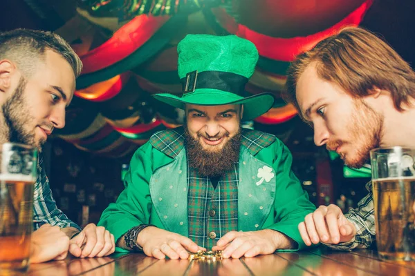 Greedy young man in green suit sit at table with friends and look at golden coins he grabbed. Other guys look at them too. Mugs of beer stand on table.