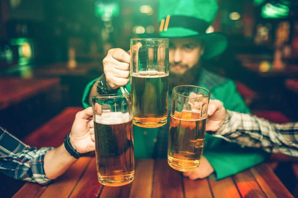 Un joven con traje verde se sienta a la mesa con amigos en el pub y mantiene juntas las jarras de cerveza. Está concentrado. . — Foto de Stock