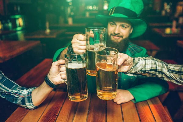 Bild eines jungen Mannes in grünem Patricks-Anzug, der mit Freunden in der Kneipe am Tisch sitzt. Sie halten Bierkrüge zusammen. — Stockfoto