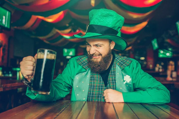 Furcsa fiatalember visel zöld St. Patricks megfeleljen. Pub asztalnál ülni, és tartsuk lenyomva a bögre barna sör. A fickó úgy néz ki, elégedett. — Stock Fotó