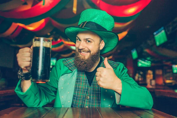 Boldog fiatalember ül tábla pub és jelentenek a kamera. Ő ragaszkodik bögre barna sör. A fickó úgy néz ki, boldog. Ő hord öltönyt St. Patricks. — Stock Fotó