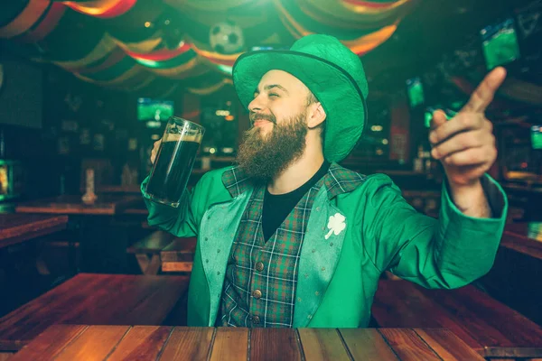 Jovem alegre feliz sente-se à mesa no pub. Ele usa fato verde de St. Patricks. Guy aponta para a frente . — Fotografia de Stock