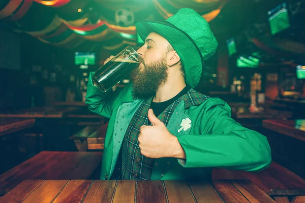Fiatal szakállas ember ül a kocsmában tábla. Ő inni sötét sör János. Fickó St. Patricks ruha hord. — Stock Fotó
