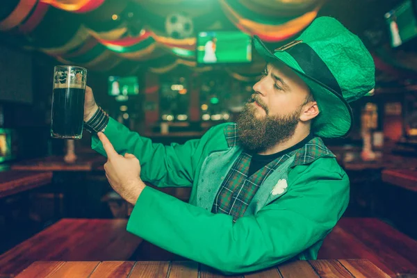 Büszke és magabiztos fiatalember kocsmában asztalnál ülni. Tartsa bögre barna sör, és mutat ez-hoz fényképezőgép. Fiatal férfi viselet St. Patricks megfeleljen. — Stock Fotó