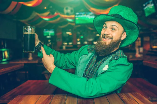O jovem positivo e alegre no terno verde senta-se à mesa no bar. Ele aponta para a caneca com cerveja escura. O tipo parece feliz. . — Fotografia de Stock