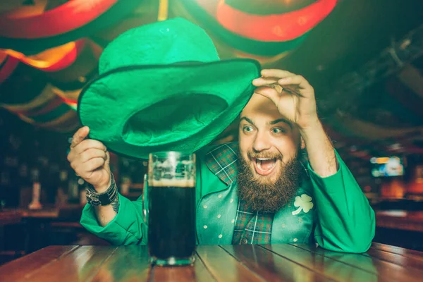 Feliz joven emocionado en traje verde sentarse a la mesa en el pub. Sostiene el sombrero encima de la taza de cerveza negra. Joven sonrisa . — Foto de Stock