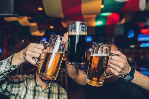 Foto de hombres jóvenes sentados en un pub y sosteniendo jarras de cerveza oscura y ligera juntos . — Foto de Stock