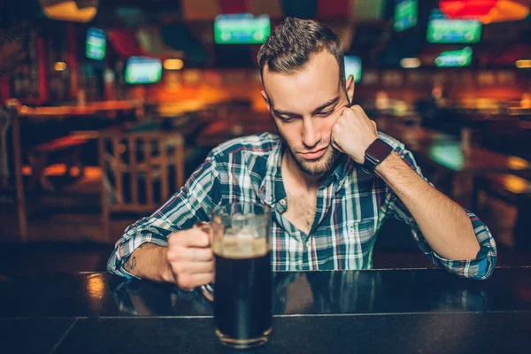 Alone young man sit at bar counter in pub. He sleep. Young man hold hand on mug with dark beer. — Stock Photo, Image