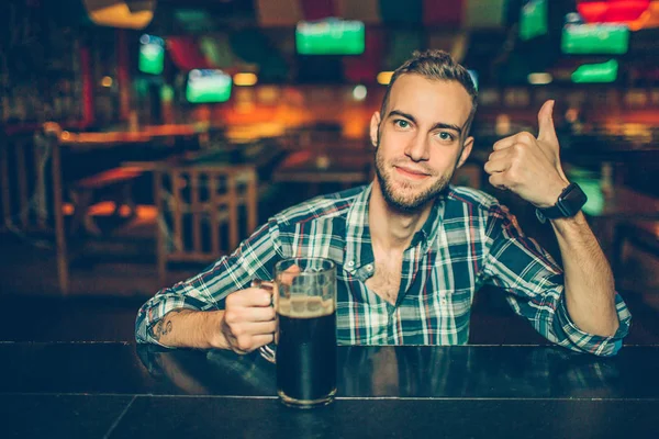 Pozitivní mladý muž sedět u baru v hospodě a podívejte se na kameru. On drží velký palec. Guy má ruku na hrnek s tmavým pivem. — Stock fotografie