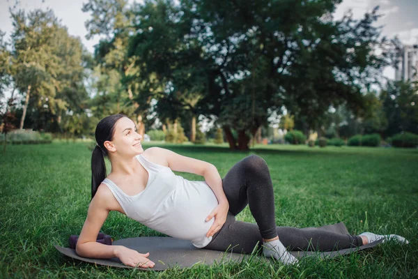 Wesoły miły młoda kobieta w ciąży leżące na jednej ręce i uśmiechając się poza w parku. Ona trzymać jeszcze jeden na brzuchu. Stoisko lewą nogę w przód z prawej. — Zdjęcie stockowe